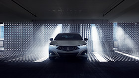 Front view of a white Acura TLX in a darkened garage with natural light shining through a gated wall behind. // Vue avant d’une Acura TLX blanche dans un garage sombre devant un mur qui laisse passer la lumière naturelle.